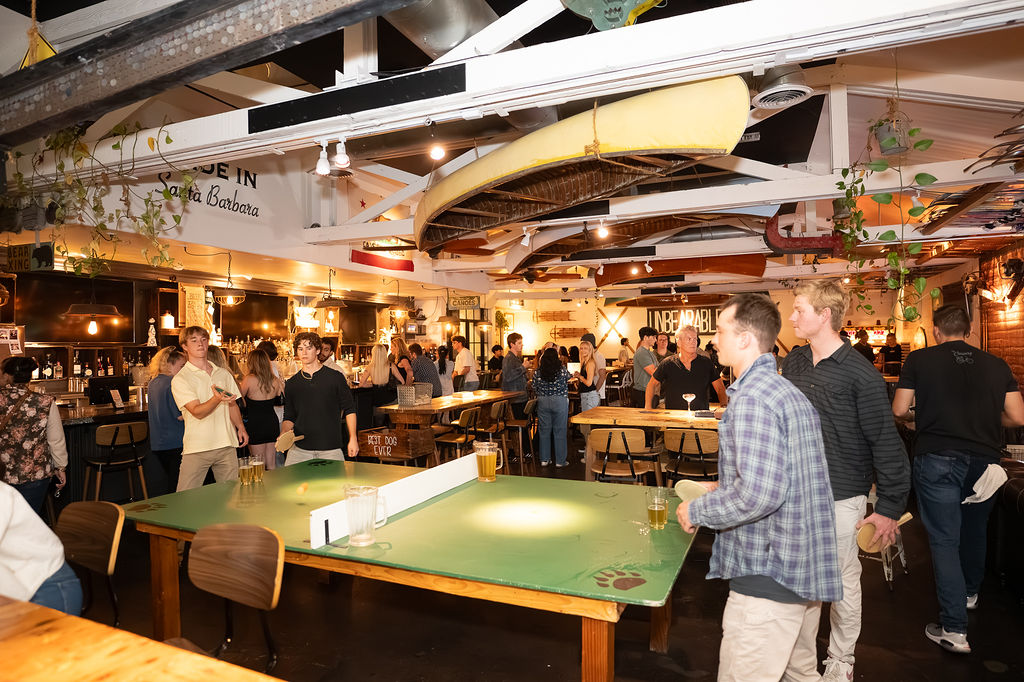 Guests playing ping pong at Unbearable Skee Lodge with a packed crowd, the UNBEARABLE sign, and canoes hanging from the ceiling